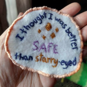 May include: A white felt patch with a peach colored border. The patch is embroidered with the text "I thought I was better than starry-eyed SAFE" in various colors.