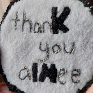 May include: A white felt circle with black stitching around the edge. The words "thank you aimee" are embroidered in silver thread with the "k" and "aimee" in black thread.