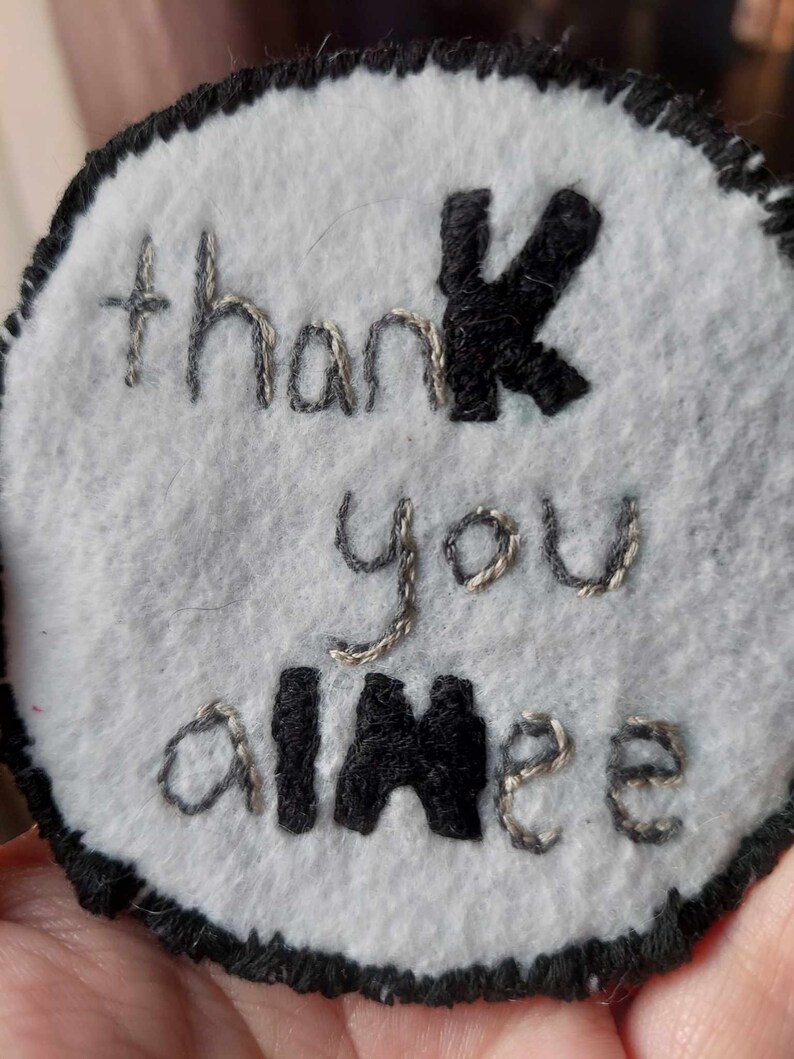 May include: A white felt circle with black stitching around the edge. The words "thank you aimee" are embroidered in silver thread with the "k" and "aimee" in black thread.