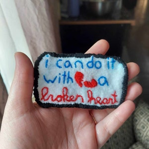 May include: A white fabric patch with black stitching around the edges. The patch has blue embroidery that reads "I can do it with a broken heart". A red embroidered heart is broken in half.
