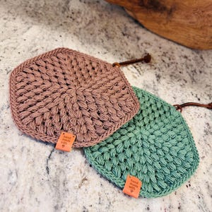 Pode incluir: Dois pegadores de panela de crochê em forma hexagonal. Um é de cor marrom claro e o outro é de cor verde claro. Ambos têm uma etiqueta de couro que diz "feito à mão com amor".