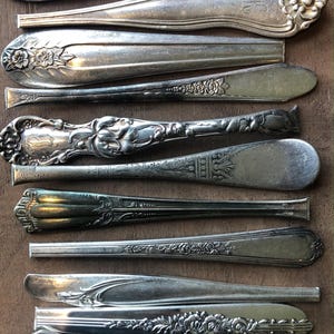 May include: An assortment of antique silverware, including spoons and a butter knife, arranged on a wooden surface. The silver utensils feature ornate floral and scrollwork designs, showcasing a vintage aesthetic. The spoons and knife are made of silver.