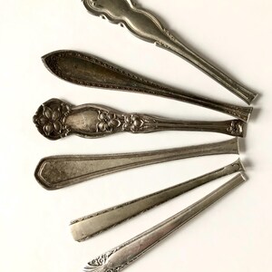 May include: An assortment of vintage silver spoons, each with unique handle designs. The spoons are arranged on a white surface, showcasing their intricate patterns and aged appearance. The spoons vary in size and style, reflecting different eras.