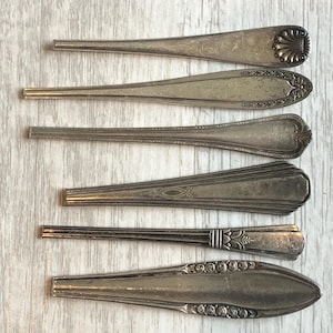 May include: Six vintage silver spoons with ornate designs are arranged on a light wood surface. The spoons vary in shape and pattern, showcasing detailed engravings and decorative elements. The spoons are likely for serving or eating.