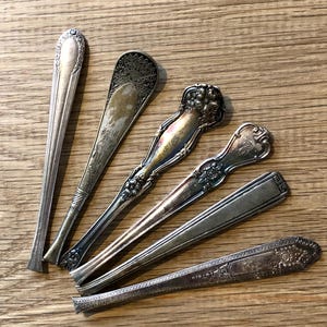 May include: A collection of six antique silver spoons with ornate designs. The spoons vary in shape and pattern, with details including floral motifs and engraved lines. They are arranged on a wooden surface, showcasing their aged appearance.