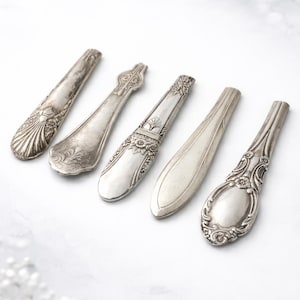 May include: An assortment of antique silver spoons with ornate designs. The spoons vary in shape and decoration, with floral and geometric patterns. The spoons are arranged on a white surface, showcasing their intricate details and vintage charm.
