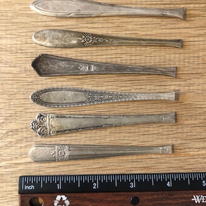 May include: Six antique silver spoons with ornate designs are arranged on a wooden surface. A ruler with inch measurements is placed below the spoons. The spoons vary in shape and decorative patterns, showcasing vintage silverware.