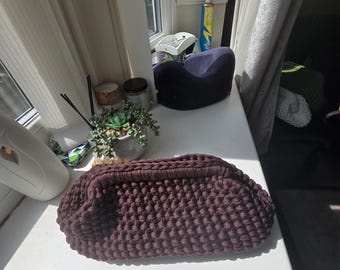May include: A dark brown, rectangular, crocheted clutch bag featuring a thick, textured yarn construction with a rigid, integrated top handle. The bag has a rounded, structured shape and a consistent, dense knot pattern throughout its exterior surface.