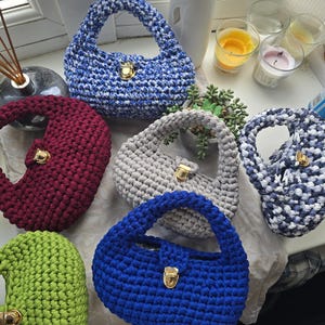 May include: Five crocheted handbags in different colors, including blue, green, gray, red, and white with blue. Each bag has a gold clasp.