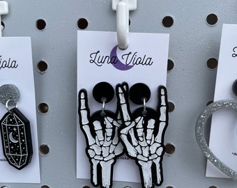 May include: A pair of black and white skeleton hand earrings with a black circle in the center. The earrings are on a white card with the text "Luna Viola" written in black.