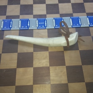 May include: A light brown antler handle with a brown leather strap tied around it. The antler handle is about 6 inches long and is lying on a wooden surface.
