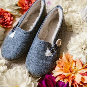 May include: A pair of gray felt slippers with white felt accents and brown button closures. The slippers are surrounded by white, orange, and purple flowers.