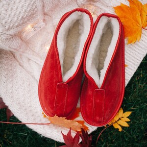 May include: A pair of red suede slippers with white fleece lining. The slippers are on a white knitted blanket with fall leaves around them.