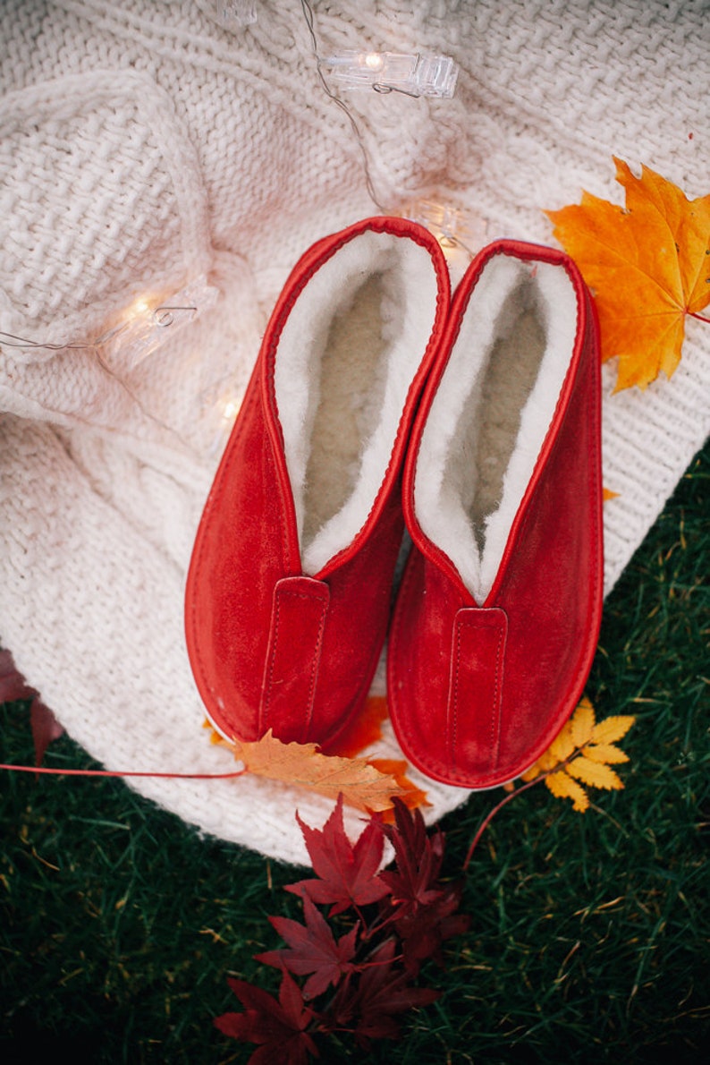 May include: A pair of red suede slippers with white fleece lining. The slippers are on a white knitted blanket with fall leaves around them.