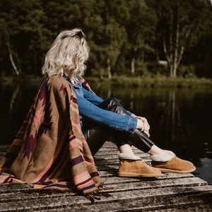 May include: A woman sits on a wooden dock wearing brown suede slippers with a white fur lining. She is wearing a brown and pink patterned blanket and blue jeans. The dock is over a body of water with green trees in the background.