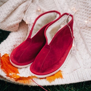 May include: A pair of red suede slippers with white soles and a fuzzy lining. The slippers are on a white knitted blanket with a string of lights.