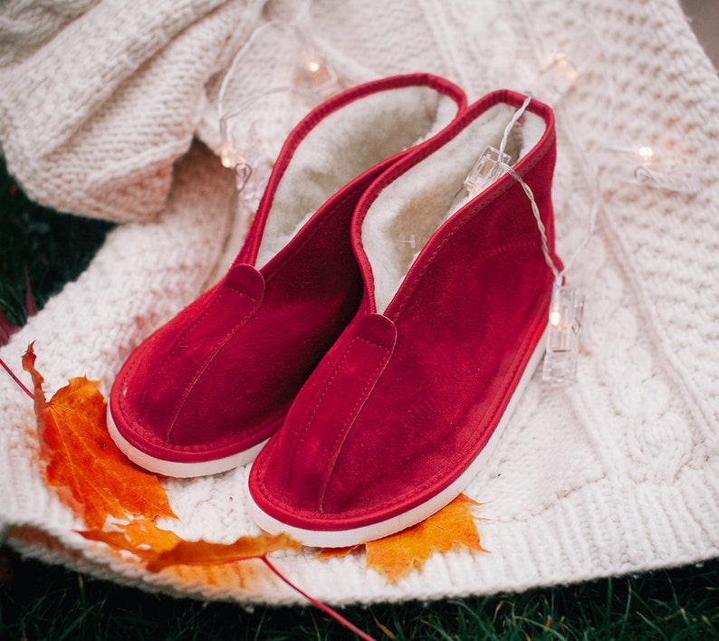 May include: A pair of red suede slippers with white soles and a fuzzy lining. The slippers are on a white knitted blanket with a string of lights.