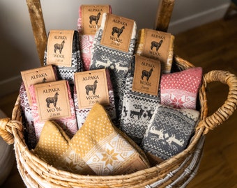 Può includere: Un cesto di vimini pieno di diversi paia di calzini di lana di alpaca. I calzini presentano vari motivi, tra cui fiocchi di neve e renne, in colori come grigio, rosso e giallo. Ogni paio ha un'etichetta con la scritta "ALPAKA WOOL".