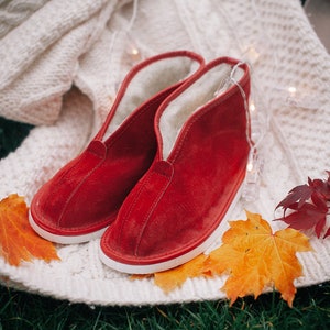 May include: A pair of red suede slippers with white soles and a fuzzy lining. The slippers are on a white knitted blanket with fall leaves and string lights.