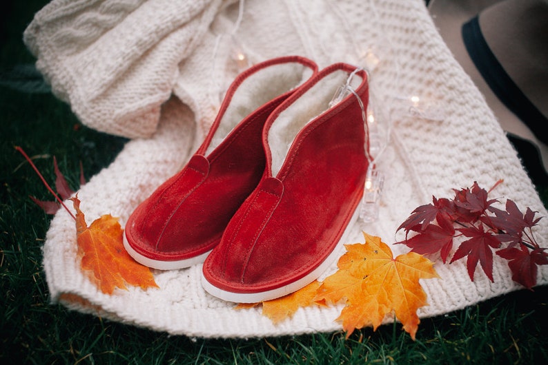 May include: A pair of red suede slippers with white soles and a fuzzy lining. The slippers are on a white knitted blanket with fall leaves and string lights.