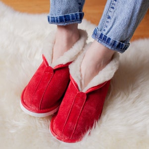 May include: Red suede slippers with white trim and a fluffy white lining. The slippers are worn on a white fluffy rug.
