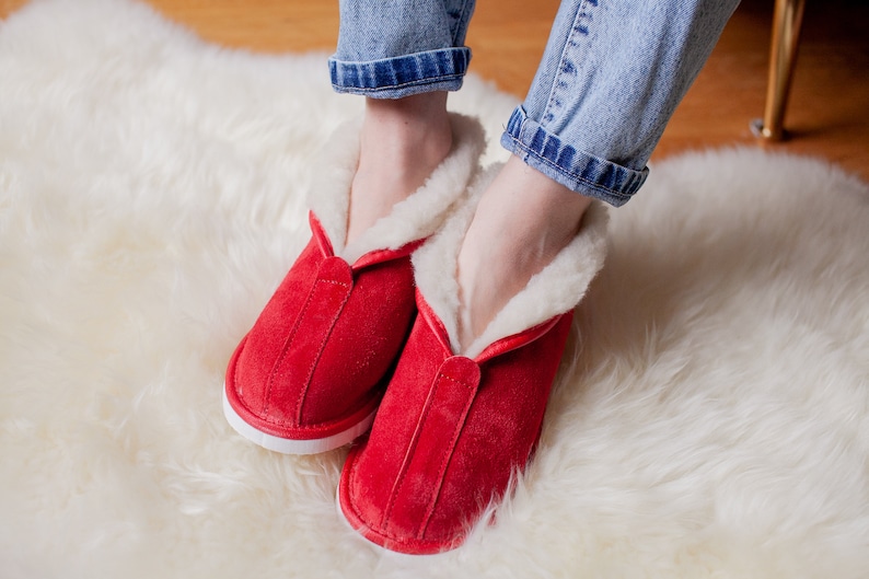 May include: Red suede slippers with white trim and a fluffy white lining. The slippers are worn on a white fluffy rug.