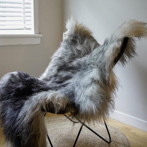 Pu&ograve; includere: Una sedia a farfalla con struttura in metallo nero e rivestimento in pelle di pecora a pelo lungo nei toni del grigio, antracite e bianco. La pelliccia spessa e strutturata copre la seduta e lo schienale, creando un aspetto morbido.