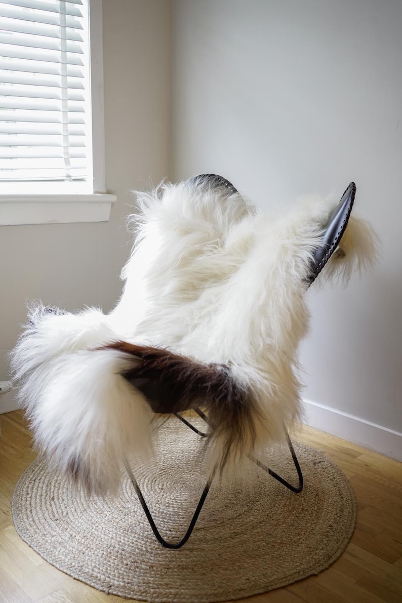 Pu&ograve; includere: Una sedia a farfalla con struttura in metallo nero e rivestimento in pelle di pecora bianca a pelo lungo con macchie marroni. La sedia presenta uno schienale curvo e una seduta profonda, con la pelliccia drappeggiata sui bordi del telaio.