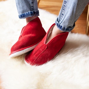 May include: A pair of red suede slippers with white soles. The slippers are worn by a person sitting on a white fluffy rug.