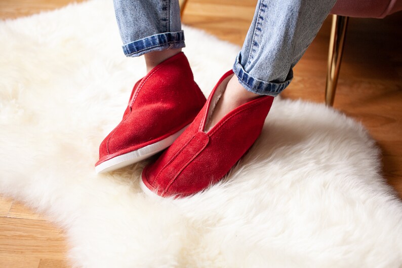 May include: A pair of red suede slippers with white soles. The slippers are worn by a person sitting on a white fluffy rug.
