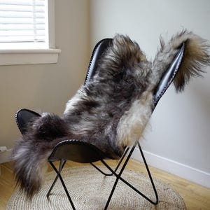 Pu&ograve; includere: Una poltrona a farfalla in pelle nera con una pelle di pecora a pelo lungo nei toni del grigio e del bianco drappeggiata sul sedile. La sedia ha una sottile struttura in metallo nero e il numero 148 &egrave; visibile in alto a sinistra.
