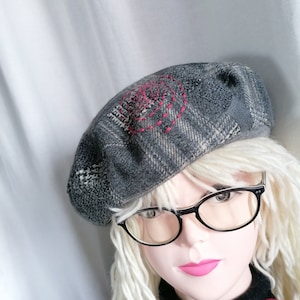 Peut inclure: Un béret en tissu gris et noir à carreaux avec un motif brodé rose. Le béret est porté sur un mannequin avec des cheveux blonds et des lunettes à monture noire.