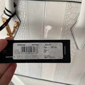 Puede incluir: Bolso de mano de cuero blanco con dise&ntilde;o a rayas y herrajes dorados. El bolso presenta un patr&oacute;n texturizado y una etiqueta con informaci&oacute;n del producto. Tiene un asa superior y una cadena dorada.