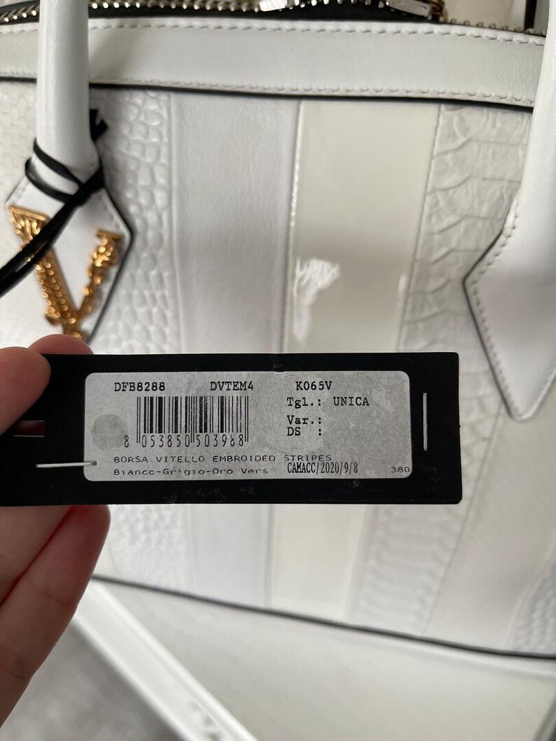 Puede incluir: Bolso de mano de cuero blanco con dise&ntilde;o a rayas y herrajes dorados. El bolso presenta un patr&oacute;n texturizado y una etiqueta con informaci&oacute;n del producto. Tiene un asa superior y una cadena dorada.