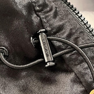 May include: A black satin-finish garment featuring a black zipper and a cylindrical metal cord lock engraved with the word VERSACE. The cord lock secures a thin black drawstring cord, and a small gold-toned circular accent is visible at the bottom.