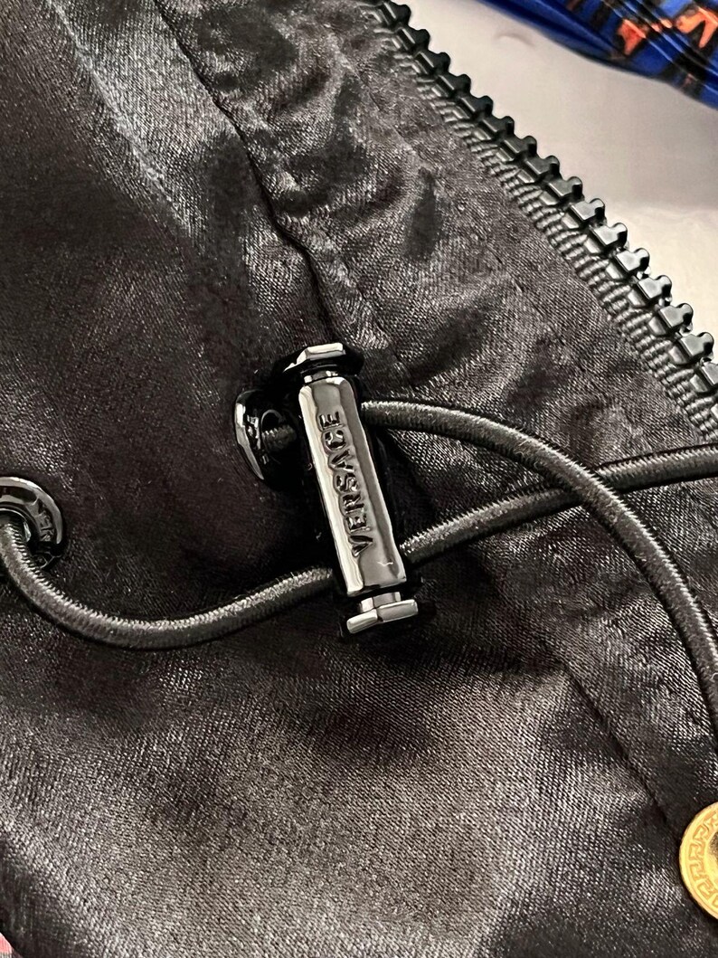 May include: A black satin-finish garment featuring a black zipper and a cylindrical metal cord lock engraved with the word VERSACE. The cord lock secures a thin black drawstring cord, and a small gold-toned circular accent is visible at the bottom.