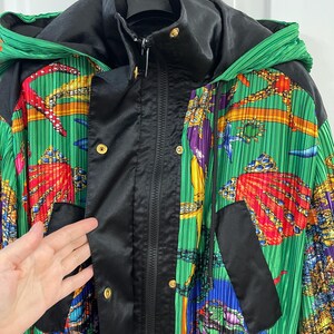May include: A hooded jacket featuring a vibrant green pleated fabric with a colorful marine-themed print of seashells and sea life. The jacket includes a black satin-finish front placket with gold-tone snap buttons and a central zipper closure.