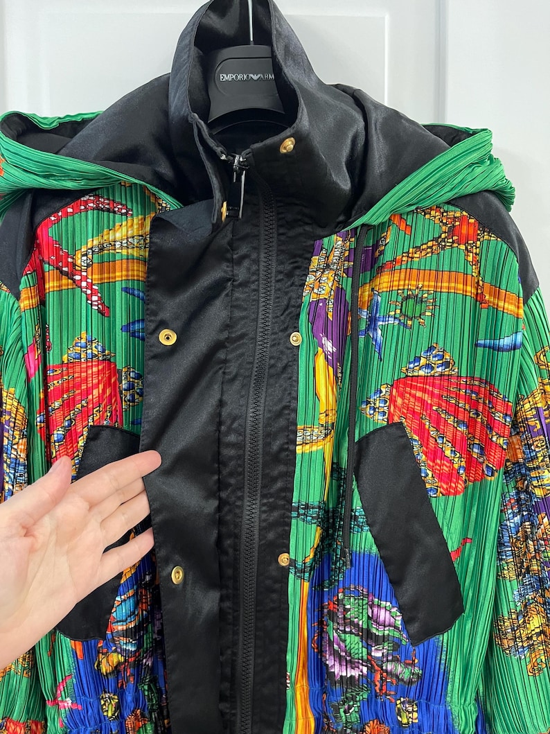 May include: A hooded jacket featuring a vibrant green pleated fabric with a colorful marine-themed print of seashells and sea life. The jacket includes a black satin-finish front placket with gold-tone snap buttons and a central zipper closure.