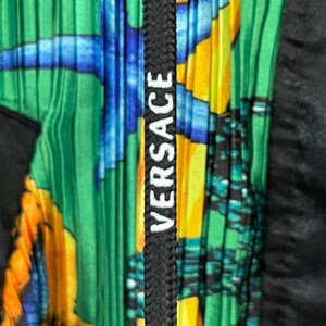 May include: A pleated garment featuring a vibrant green, yellow, and blue aquatic-themed print. A black drawstring cord runs vertically down the center, displaying the white text VERSACE printed in a bold, sans-serif font along its length.