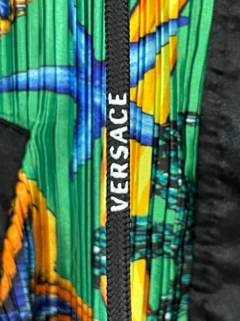 May include: A pleated garment featuring a vibrant green, yellow, and blue aquatic-themed print. A black drawstring cord runs vertically down the center, displaying the white text VERSACE printed in a bold, sans-serif font along its length.
