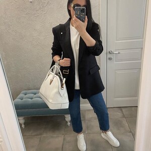 Puede incluir: Una persona con una blazer negra, camiseta blanca y vaqueros azules, sosteniendo un bolso blanco. La persona tambi&eacute;n lleva zapatillas blancas y gafas de sol. El fondo incluye un espejo, una l&aacute;mpara y un banco azul.