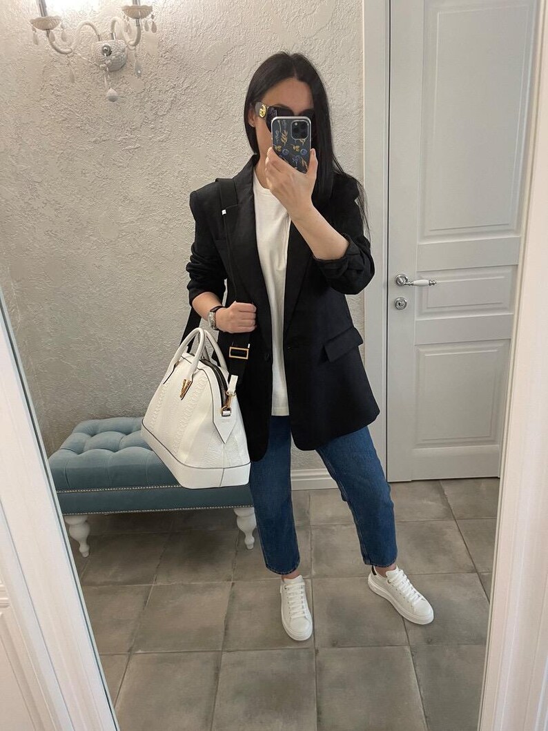 Puede incluir: Una persona con una blazer negra, camiseta blanca y vaqueros azules, sosteniendo un bolso blanco. La persona tambi&eacute;n lleva zapatillas blancas y gafas de sol. El fondo incluye un espejo, una l&aacute;mpara y un banco azul.
