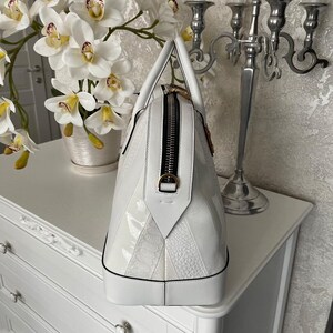 Puede incluir: Un bolso de mano de cuero blanco con forma de c&uacute;pula estructurada. El bolso tiene una cremallera negra, herrajes dorados y una mezcla de paneles lisos y texturizados. El bolso est&aacute; sobre una c&oacute;moda blanca.