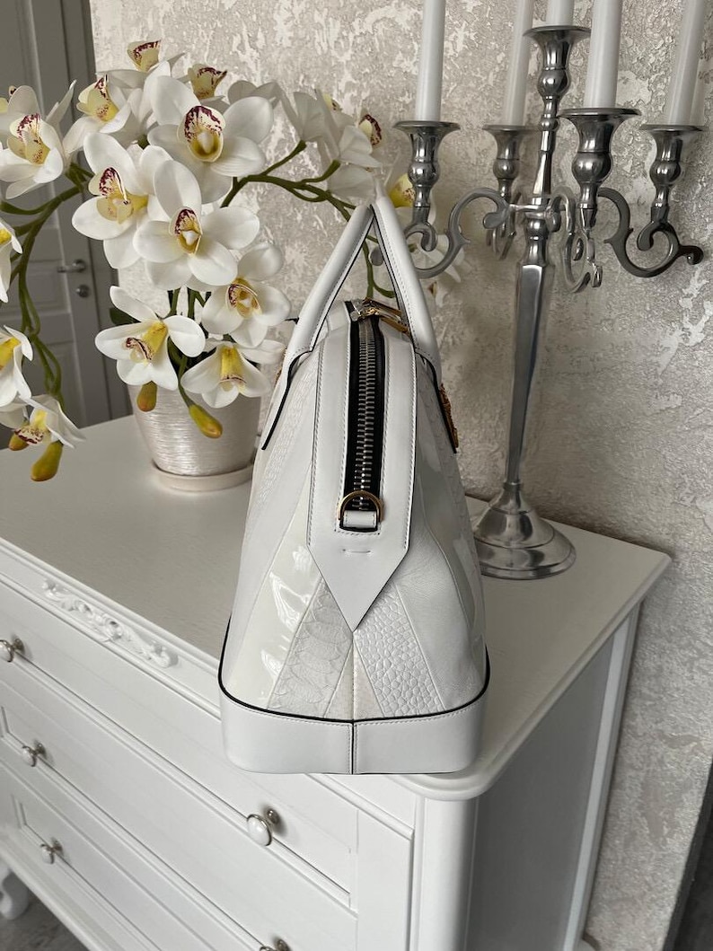 Puede incluir: Un bolso de mano de cuero blanco con forma de c&uacute;pula estructurada. El bolso tiene una cremallera negra, herrajes dorados y una mezcla de paneles lisos y texturizados. El bolso est&aacute; sobre una c&oacute;moda blanca.
