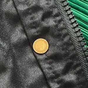 May include: A black fabric garment featuring a gold-toned circular button with a Greek key pattern embossed around the edge. The item includes a black plastic zipper track running vertically along the side of the buttoned placket.