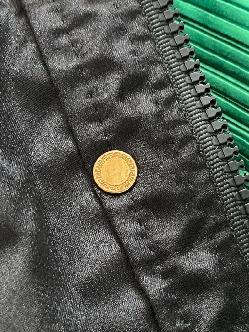 May include: A black fabric garment featuring a gold-toned circular button with a Greek key pattern embossed around the edge. The item includes a black plastic zipper track running vertically along the side of the buttoned placket.