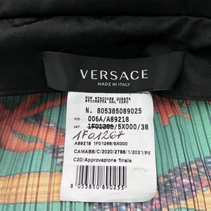 May include: A black Versace garment label with white text reading MADE IN ITALY, attached to a white care tag featuring the text N. 805385089025, MOD. 006A/A89218, ART. 1F01266/5X000/38, a barcode, and a colorful pleated fabric background with orange and green.