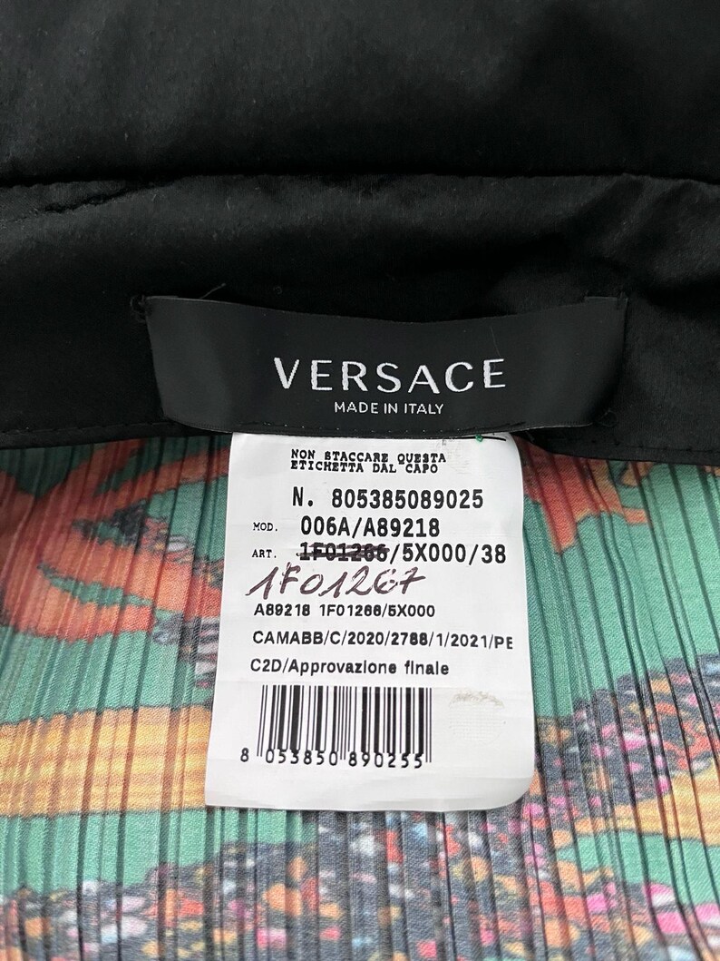 May include: A black Versace garment label with white text reading MADE IN ITALY, attached to a white care tag featuring the text N. 805385089025, MOD. 006A/A89218, ART. 1F01266/5X000/38, a barcode, and a colorful pleated fabric background with orange and green.