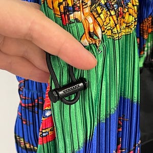 May include: A pleated garment featuring a vibrant, multicolored graphic print in shades of blue, green, yellow, and red. A black cord lock with the word VERSACE embossed in white is attached to a black drawstring, cinching the pleated fabric.