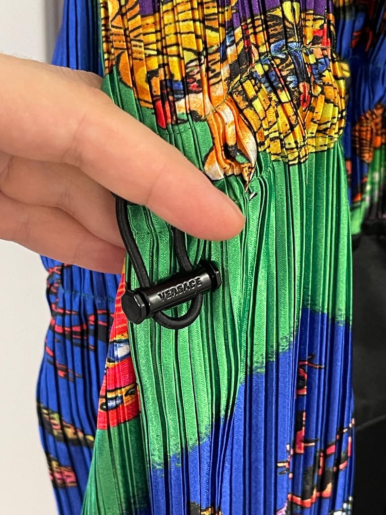 May include: A pleated garment featuring a vibrant, multicolored graphic print in shades of blue, green, yellow, and red. A black cord lock with the word VERSACE embossed in white is attached to a black drawstring, cinching the pleated fabric.